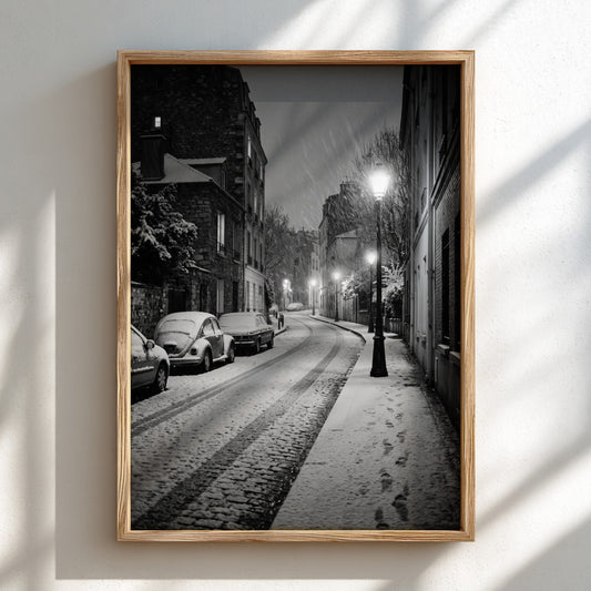 Black and white poster of empty city street during snowfall with parked cars and glowing street lamps
