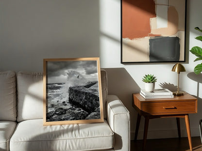 Black and white poster of waves crashing against rugged stone wall under cloudy sky with seagull flying
