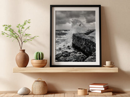 Black and white poster of waves crashing against rugged stone wall under cloudy sky with seagull flying